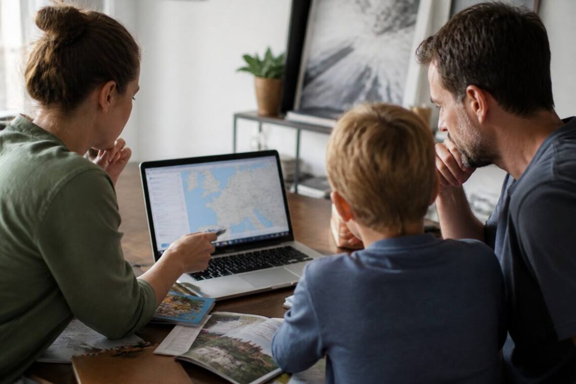 Gezin dat aan een vakantietafel reisopties vergelijkt op een laptop en reisgidsen bekijkt